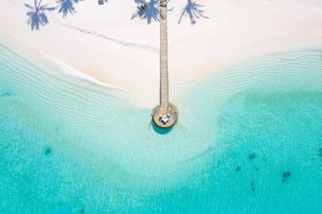 Luftaufnahme eines Holzstegs, der zu einer Plattform im türkisfarbenen Wasser der Malediven führt. Weißer Sandstrand und Palmen. Maledives Special Benefit.