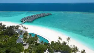 Luftaufnahme von Raaya by Atmosphere: Überwasserbungalows auf türkisfarbenem Wasser, weißer Sandstrand, üppige Vegetation und ein einladender Pool. Paradiesische Malediven-Szene.