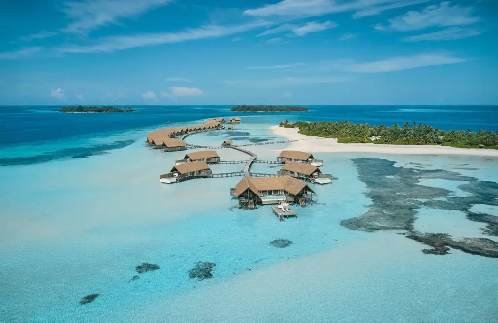 Luftaufnahme von Como Cocoa: Luxuriöse Wasserbungalows auf türkisfarbenem Wasser, verbunden durch Holzstege, mit tropischen Inseln im Hintergrund.