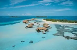 Luftaufnahme von Como Cocoa: Luxuriöse Wasserbungalows auf türkisfarbenem Wasser, verbunden durch Holzstege, mit tropischen Inseln im Hintergrund.