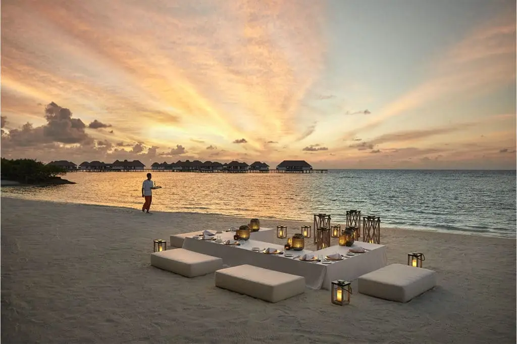 Romantisches Abendessen am Strand im Como Maalifushi bei Sonnenuntergang. Ein Kellner serviert, während die Sonne den Himmel in goldenen Farben malt. Luxuriöse Wasserbungalows im Hintergrund.