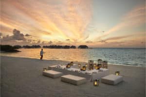 Romantisches Abendessen am Strand im Como Maalifushi bei Sonnenuntergang. Ein Kellner serviert, während die Sonne den Himmel in goldenen Farben malt. Luxuriöse Wasserbungalows im Hintergrund.