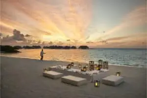 Romantisches Abendessen am Strand im Como Maalifushi bei Sonnenuntergang. Ein Kellner serviert, während die Sonne den Himmel in goldenen Farben malt. Luxuriöse Wasserbungalows im Hintergrund.