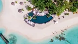Luftaufnahme von Constance Moofushi: Türkisblaues Wasser umspült einen weißen Sandstrand mit Pool, Liegen und Palmen. Ein Herz ist in den Sand gemalt. Paradiesische Urlaubsstimmung.