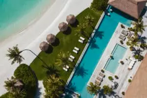 Luftaufnahme von Ithaafushi Private Island: Türkisfarbenes Wasser, weißer Sandstrand, grüne Rasenfläche mit Sonnenschirmen und Liegen, sowie ein luxuriöser Poolbereich mit Palmen.