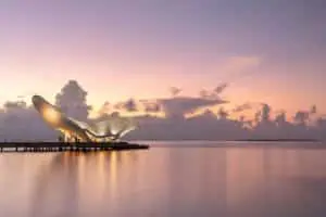 Joali Being: Atemberaubendes Gebäude im futuristischen Design auf einem Steg im Wasser bei Sonnenuntergang. Der Himmel ist in sanfte Rosa- und Lilatöne getaucht.