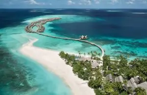 Luftaufnahme von Joali Maldives: Luxuriöse Wasserbungalows auf türkisfarbenem Wasser, verbunden durch einen Holzsteg zu einem Strand mit üppiger Vegetation.