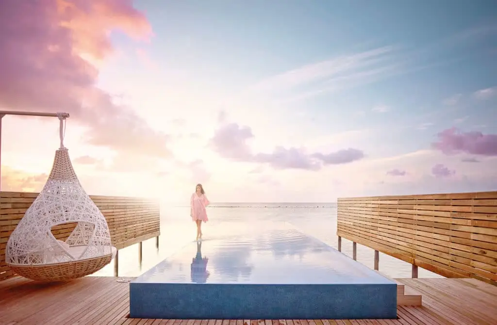 Frau im rosa Kleid spaziert über den Infinity-Pool im Lux South Ari Atoll bei Sonnenuntergang. Hängesessel und Holzdeck ergänzen die tropische Szene.