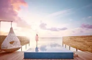 Frau im rosa Kleid spaziert über den Infinity-Pool im Lux South Ari Atoll bei Sonnenuntergang. Hängesessel und Holzdeck ergänzen die tropische Szene.