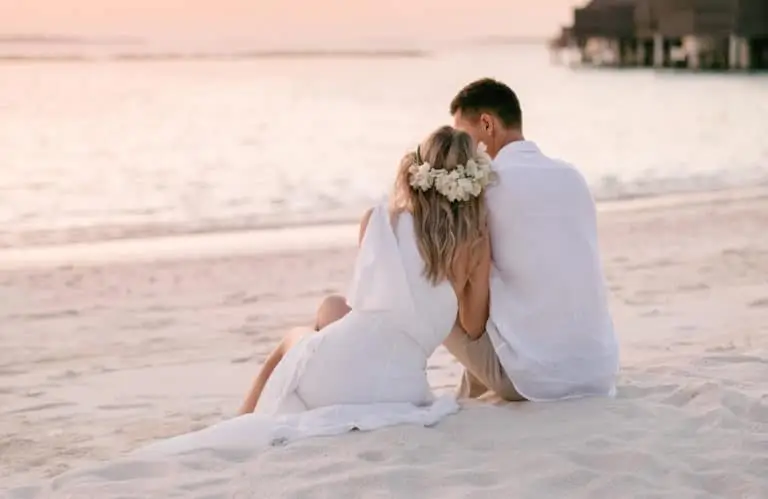Ein Paar sitzt am Strand der Malediven, die Frau trägt ein weißes Kleid und einen Blumenkranz. Romantische Szene für eine Hochzeit auf den Malediven bei Sonnenuntergang.