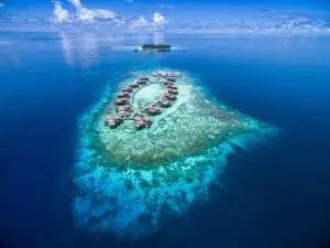 Luftaufnahme von Raffles Maldives Meradhoo: Luxuriöse Wasserbungalows auf einer tropischen Insel, umgeben von türkisfarbenem Wasser und einem fernen Inselchen unter blauem Himmel.