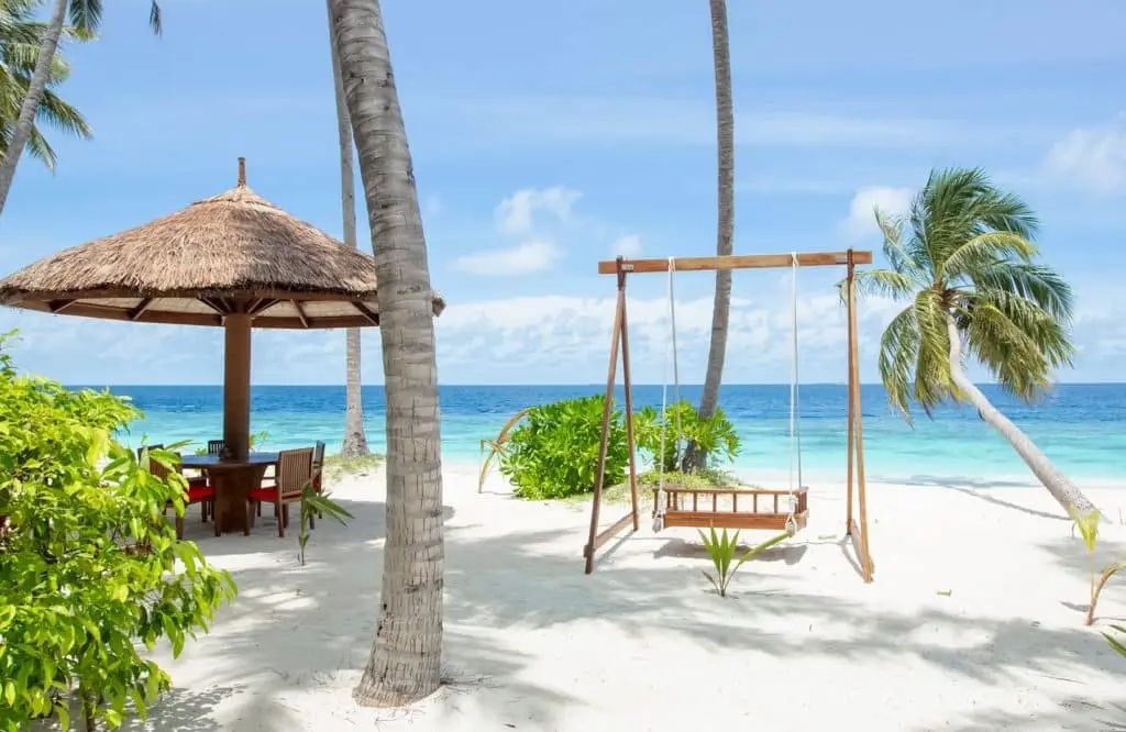 Idyllische Szene im Reethi Faru: Schaukel am weißen Sandstrand, ein Strohdach-Pavillon mit Tisch und Stühlen, türkisfarbenes Meer und Palmen unter blauem Himmel.