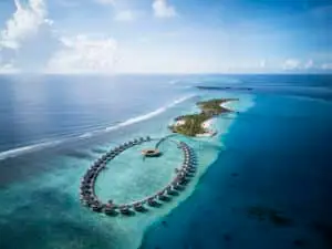 Luftaufnahme des Ritz-Carlton Maldives: Luxuriöse Wasserbungalows in einem perfekten Kreis angeordnet, türkisfarbenes Wasser und tropische Inseln im Hintergrund. Ein Paradies.