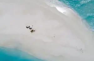 Luftaufnahme von Soneva Fushi: Weißer Sandstrand, türkisfarbenes Wasser, zwei Sonnenschirme und Personen auf einer Decke, die die friedliche Atmosphäre des Luxusresorts genießen.