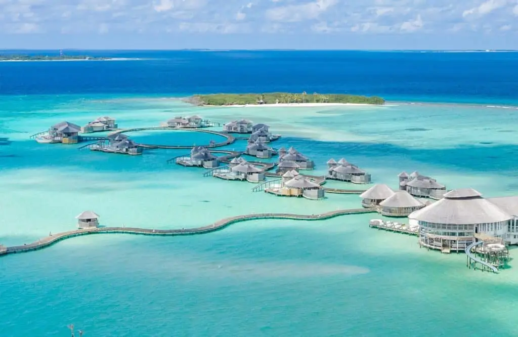 Luxuriöse Wasserbungalows von Soneva Jani, Malediven. Türkisblaues Wasser umgibt die strohgedeckten Villen, verbunden durch Holzstege. Im Hintergrund eine tropische Insel unter blauem Himmel.