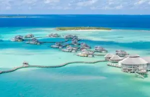 Luxuriöse Wasserbungalows von Soneva Jani, Malediven. Türkisblaues Wasser umgibt die strohgedeckten Villen, verbunden durch Holzstege. Im Hintergrund eine tropische Insel unter blauem Himmel.