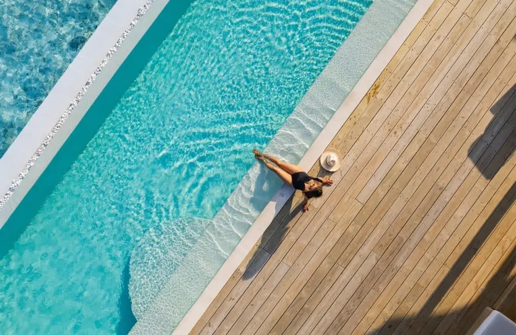 Luftaufnahme von einer Frau im schwarzen Badeanzug, die am Pool des Soneva Jani Resorts auf einem Holzdeck entspannt. Ein Strohhut liegt daneben.