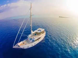 Die Soneva in Aqua, eine luxuriöse Yacht, gleitet auf tiefblauem Wasser an einer kleinen Insel vorbei. Der strahlend weiße Rumpf und das helle Deck kontrastieren mit dem Ozean.