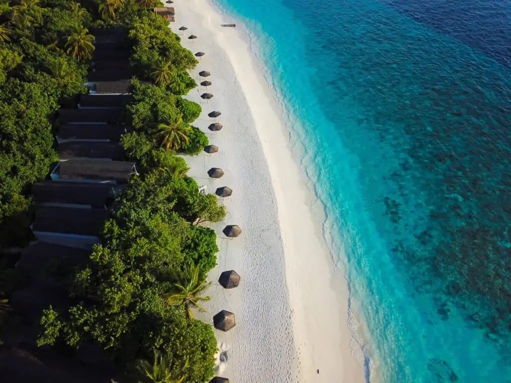 Luftaufnahme von Reethi Beach mit türkisfarbenem Wasser, weißem Sandstrand, Strohschirmen und Villen im NH Reethi Resort, umgeben von üppigem Grün.