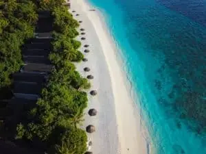 Luftaufnahme von Reethi Beach mit türkisfarbenem Wasser, weißem Sandstrand, Strohschirmen und Villen im NH Reethi Resort, umgeben von üppigem Grün.