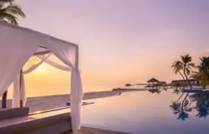 Sonnenuntergang im Nooe Kunaavashi Resort Malediven: Ein luxuriöser Infinity-Pool spiegelt den rosafarbenen Himmel, mit einem weißen Baldachin im Vordergrund und Palmen am Horizont.