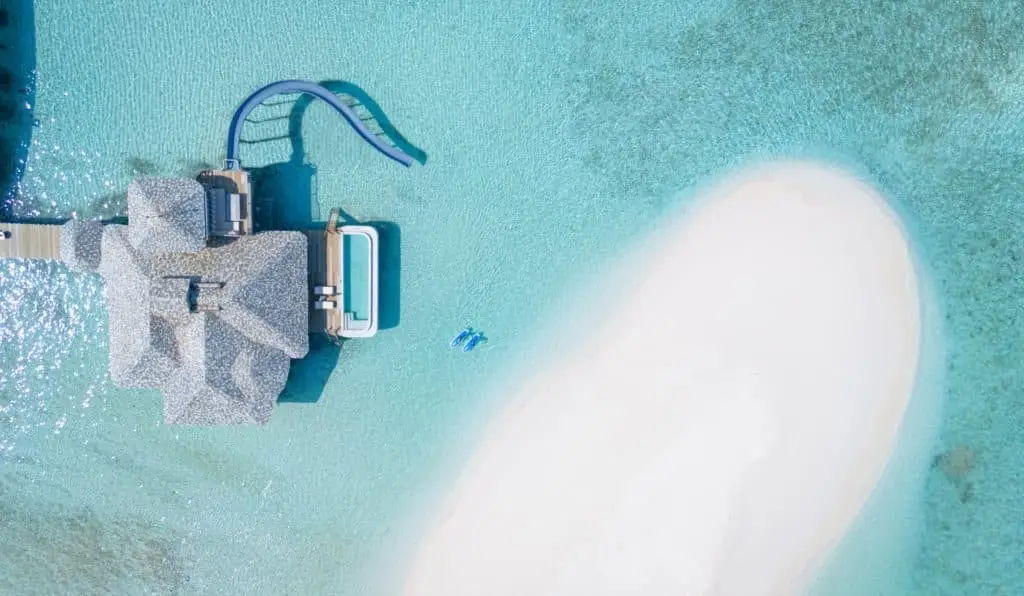 Luftaufnahme von Soneva Secret: Luxusvilla mit Wasserrutsche, Pool und Sandbank im türkisfarbenen Wasser. Zwei blaue Schwimmkörper treiben im Meer.