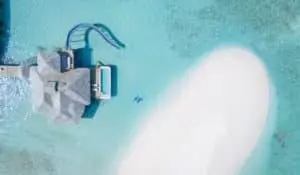 Luftaufnahme von Soneva Secret: Luxusvilla mit Wasserrutsche, Pool und Sandbank im türkisfarbenen Wasser. Zwei blaue Schwimmkörper treiben im Meer.