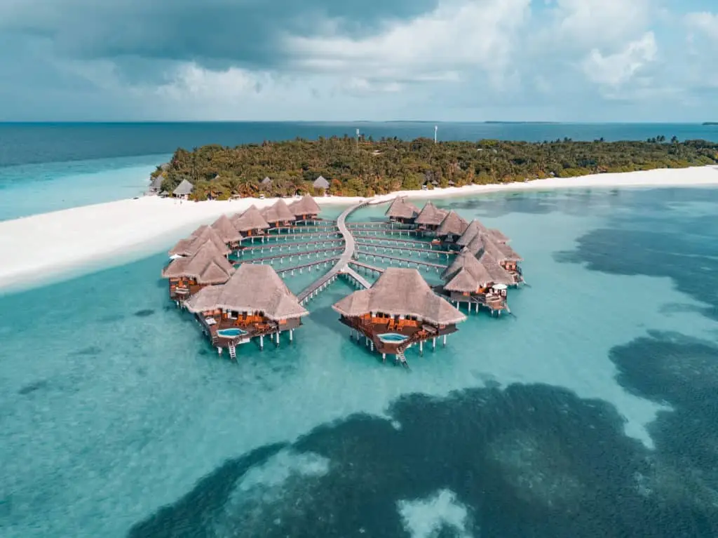 Luftaufnahme von Luxus-Wasserbungalows mit Strohdächern im Coco Palm Dhuni Kolhu Resort, Malediven. Türkisfarbenes Wasser umgibt die Anlage.