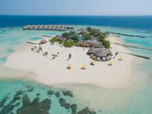 Luftaufnahme des Drift Thelu Veliga Retreat: Eine tropische Insel mit strohgedeckten Hütten, weißen Sandstränden und türkisfarbenem Wasser. Im Hintergrund Wasserbungalows auf Stelzen.