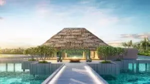 Luxuriöses InterContinental Maamunagau Resort: Strohgedecktes Gebäude auf Stelzen über türkisfarbenem Wasser, verbunden durch einen Holzsteg. Tropische Bäume und strahlend blauer Himmel.