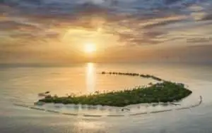 Luftaufnahme des JW Marriott Maldives Resort bei Sonnenaufgang. Eine tropische Insel mit Villen über dem Wasser, umgeben von türkisfarbenem Wasser und einem goldenen Himmel.
