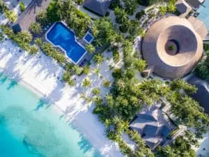 Luftaufnahme von Meeru Island: Türkisfarbenes Wasser trifft auf weißen Sandstrand, gesäumt von Palmen. Ein Pool und ein strohgedecktes Gebäude runden das tropische Paradies ab.