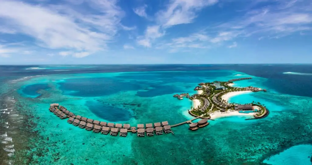 Luftaufnahme des Hilton Amingiri Malediven Resorts. Türkisfarbenes Wasser umgibt die Insel mit Wasserbungalows und weißen Sandstränden unter blauem Himmel.