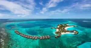 Luftaufnahme des Hilton Amingiri Malediven Resorts. Türkisfarbenes Wasser umgibt die Insel mit Wasserbungalows und weißen Sandstränden unter blauem Himmel.
