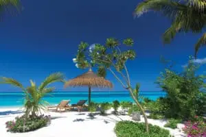 Tropischer Strand im Atmosphere Kanifushi: Liegestühle unter einem Strohschirm, türkisfarbenes Meer und weißer Sandstrand unter strahlend blauem Himmel.