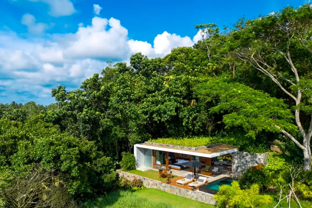 Luftaufnahme einer luxuriösen HARITHA Villa, eingebettet in üppiges Grün unter blauem Himmel. Pool, Sonnenterrasse und Dachgarten verschmelzen mit der Natur.