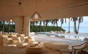 Innenansicht des Luxus-Resorts AMAN Malediven: Elegante Lounge mit Blick auf Pool, Strand und Palmen. Helle Möbel und sanfte Beleuchtung schaffen eine entspannte Atmosphäre.