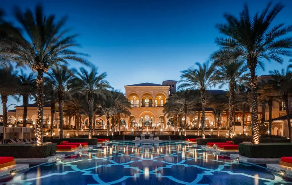 Exklusives Strandhotel Dubai: Das One & Only The Palm bei Nacht. Beleuchteter Pool spiegelt Palmen und die Fassade des Hotels wider, unter tiefblauem Himmel.
