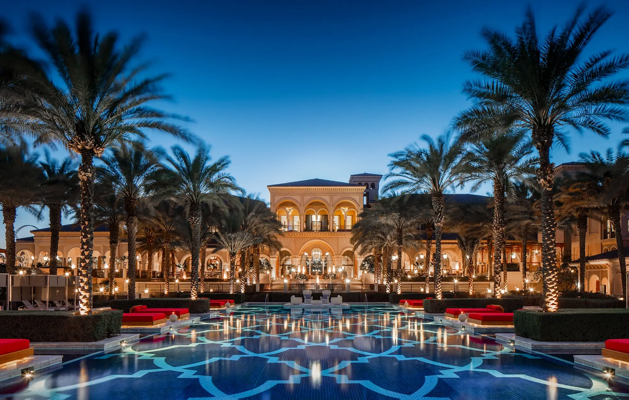 Exklusives Strandhotel Dubai: Das One & Only The Palm bei Nacht. Beleuchteter Pool spiegelt Palmen und die Fassade des Hotels wider, unter tiefblauem Himmel.