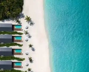 Luftaufnahme eines Boutique Luxusresorts auf den Malediven: Türkisblaues Wasser trifft auf weißen Sandstrand mit privaten Villen und Pools. Palmen spenden Schatten, ein idyllisches Urlaubsparadies.