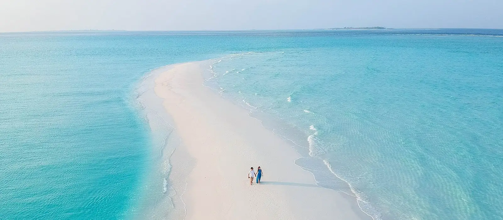 Paar spaziert Hand in Hand auf einer strahlend weißen Sandbank auf den Malediven. Türkisblaues Wasser umgibt die Insel. Ideale Reisezeit für die Malediven.