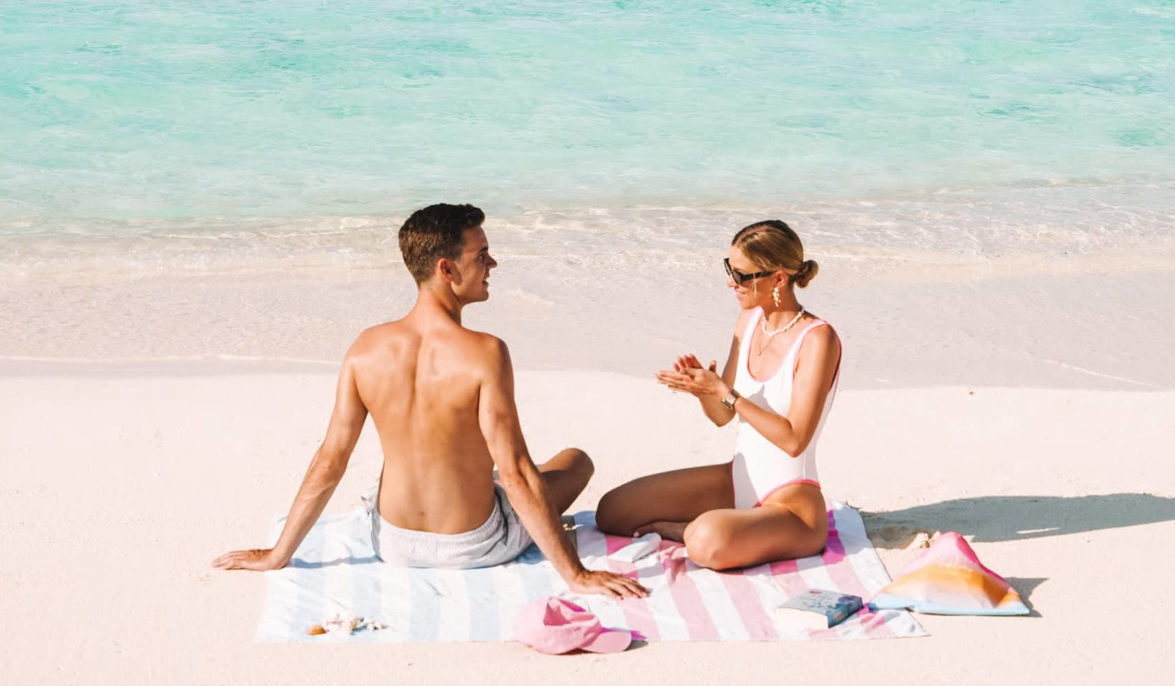 Ein Paar sitzt am weißen Sandstrand vor türkisfarbenem Wasser. Sie tragen Badebekleidung, eine Sonnenbrille und Schmuck. @betterexploretogether genießt die Sonne am Meer.