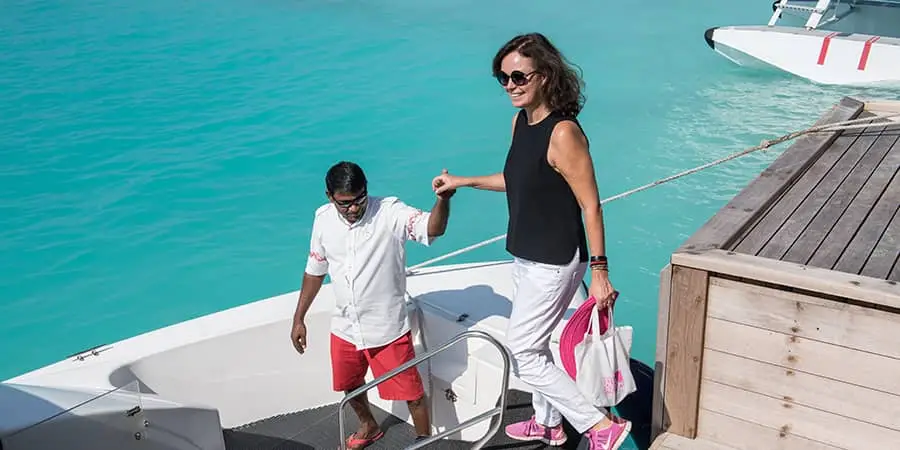 Frau steigt mit Hilfe eines Angestellten von einem Boot auf einen Steg auf den Malediven. Türkisblaues Wasser und ein strahlender Tag. Inselhüpfen im Paradies.