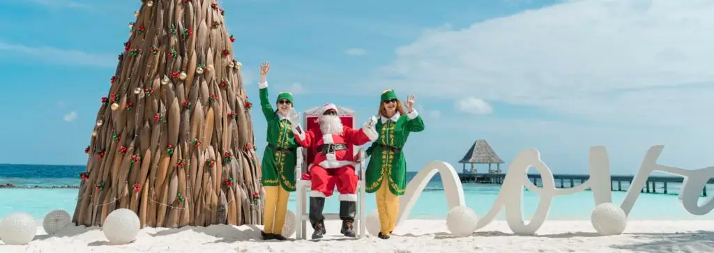 Weihnachtliche Szene im Nova Maldives: Santa sitzt mit zwei Elfen vor einem ungewöhnlichen Weihnachtsbaum aus Holz am Strand. Fröhliche Feiertage im Paradies!