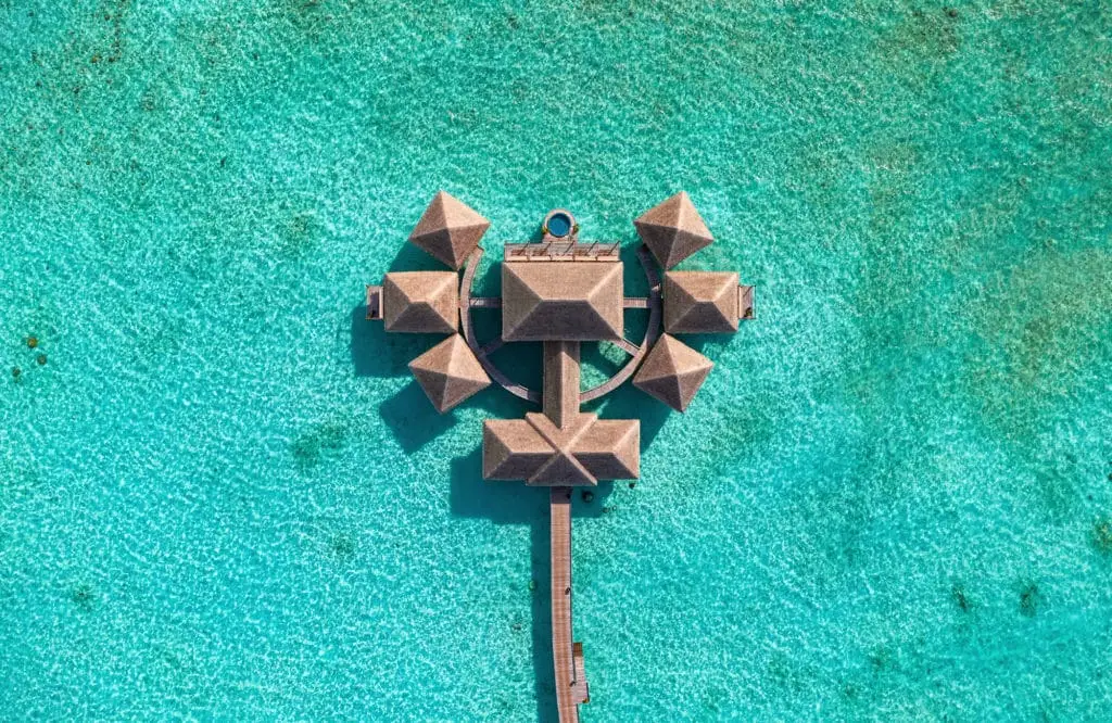 Vogelperspektive auf das Velaa Underwater Spa, ein luxuriöses Resort mit strohgedeckten Hütten, verbunden durch Stege über türkisfarbenem Wasser. Ein unvergessliches Erlebnis.