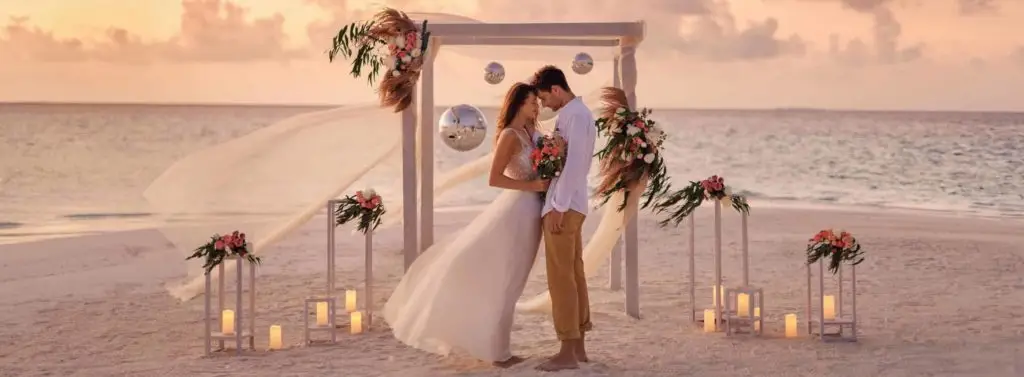 Barfußhochzeit im Paradies: Ein verliebtes Paar steht unter einem dekorierten Traubogen am Strand von Nova Maldives bei Sonnenuntergang. Kerzenlicht und Blumenschmuck schaffen eine romantische Atmosphäre.