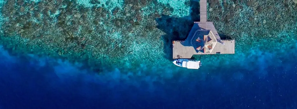 Luftaufnahme eines luxuriösen Holzstegs mit Pavillon im türkisfarbenen Wasser des Banyan Tree Vabbinfaru. Ein Boot liegt am Steg vor Anker.