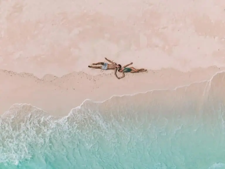 Luftaufnahme eines Paares, das Händchen haltend im Sand am Strand liegt, mit türkisfarbenem Wasser im Hintergrund.