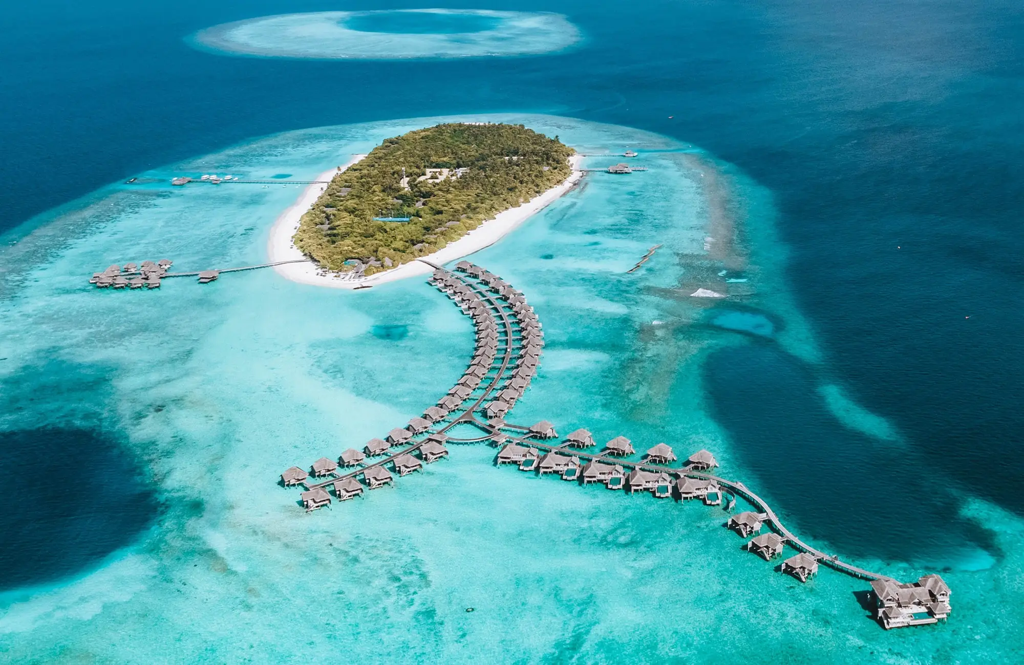 Luftaufnahme einer tropischen Insel mit Resort und über Stege verbundenen Wasserbungalows im türkisfarbenen Meer auf den Malediven