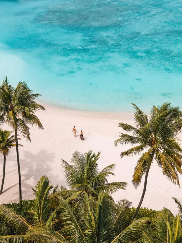 Zwei Personen gehen an einem weißen Sandstrand zwischen Palmen am türkisfarbenen Meer entlang